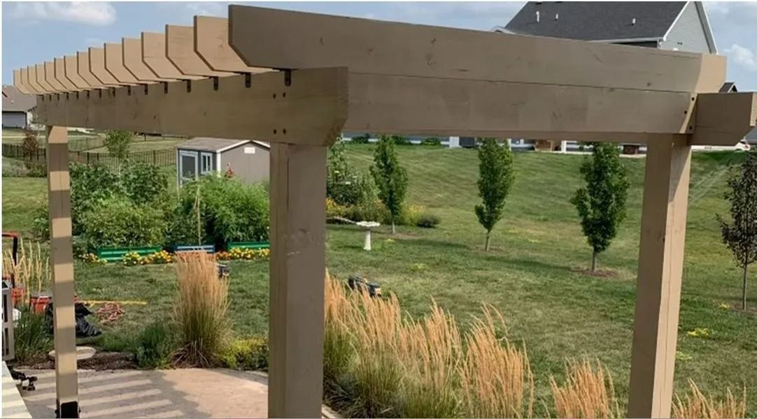 Tan pergola in a yard with green grass, plants, and small trees.