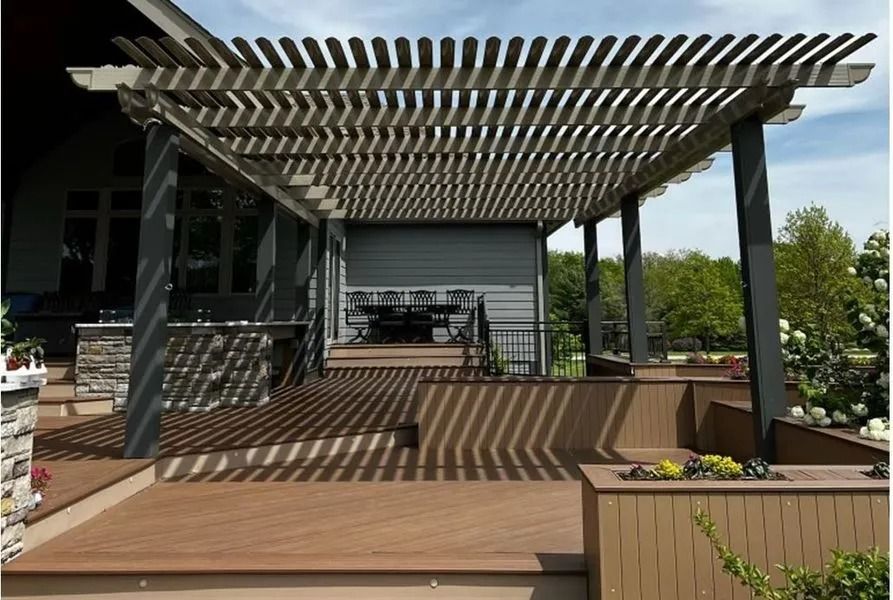 Wooden pergola over a deck, casting shadows. Brown deck and planters, with a house in the background.