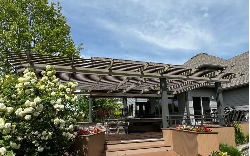 Wooden pergola over a deck, house in the background, blue sky with clouds.
