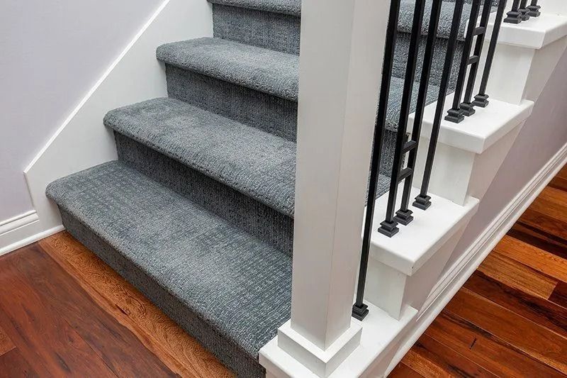 Gray carpeted stairs with white trim and black metal railing, adjacent to hardwood flooring.