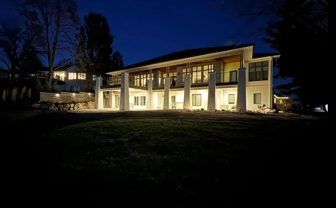 Two-story house at dusk, lit from within, with long colonnade. Lawn in foreground, trees in background.