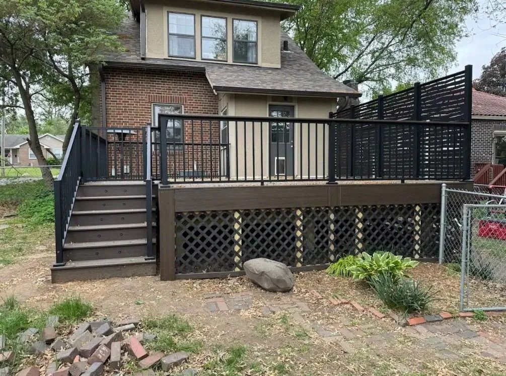 Backyard deck with dark stairs, black railings, and decorative lattice skirting. House with brick facade visible.