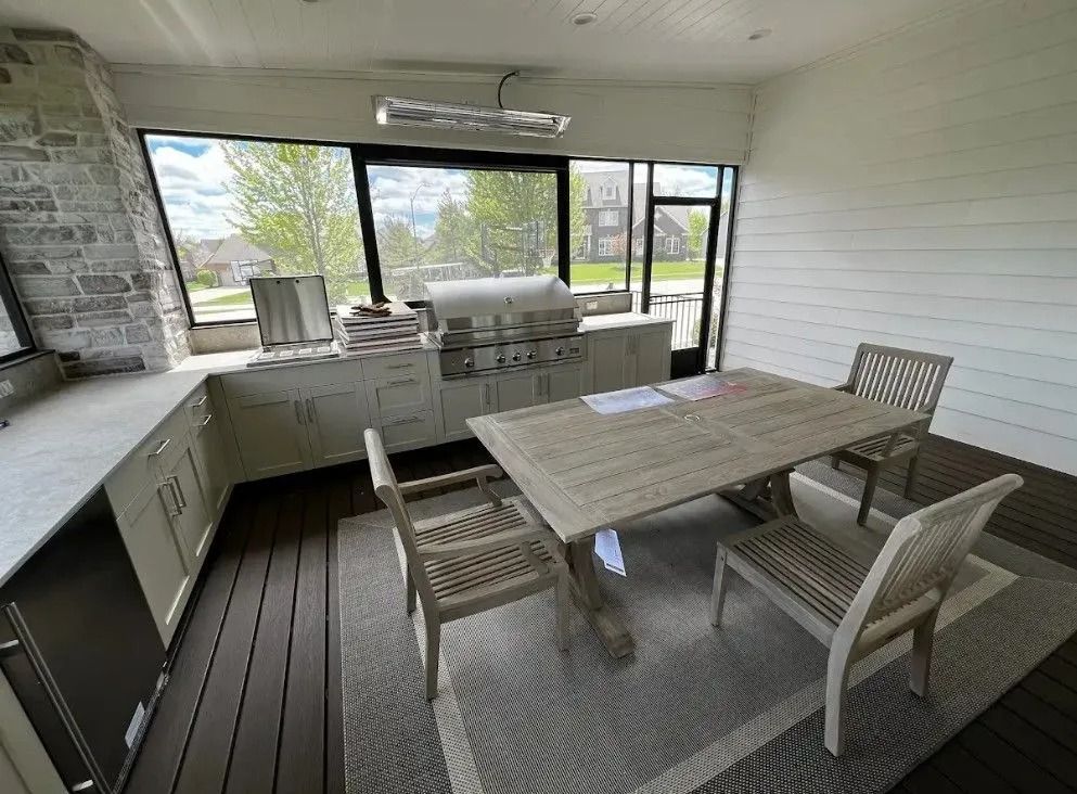 Outdoor kitchen with built-in grill, cabinets, and dining table on a deck with windows, surrounded by trees.