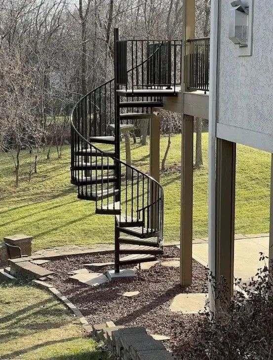 Black spiral staircase descends from a deck to a grassy yard with trees in the background.
