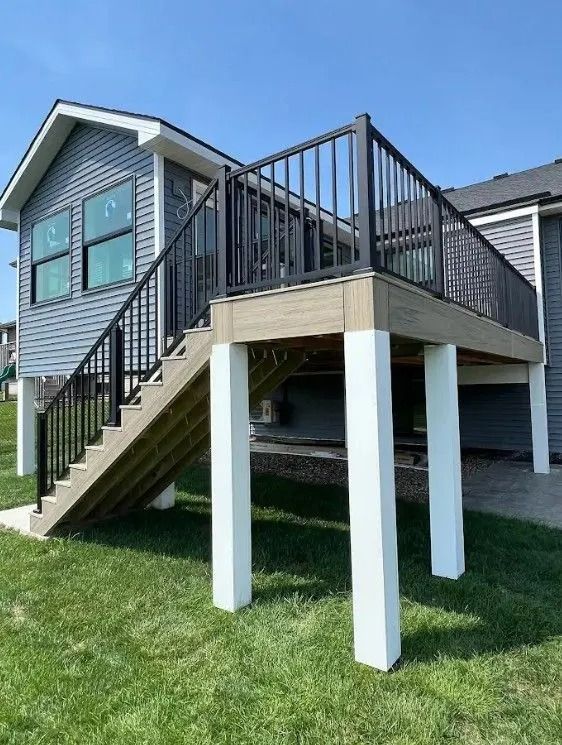 Elevated wooden deck with black railings and white support columns attached to a gray house; green grass below.