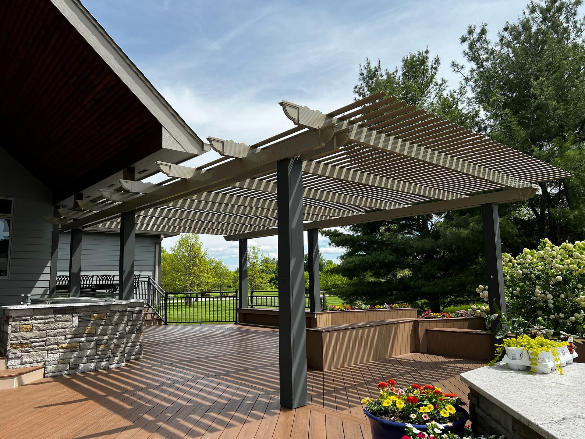 Backyard patio with striped pergola, stone planter, and flower beds beside a house