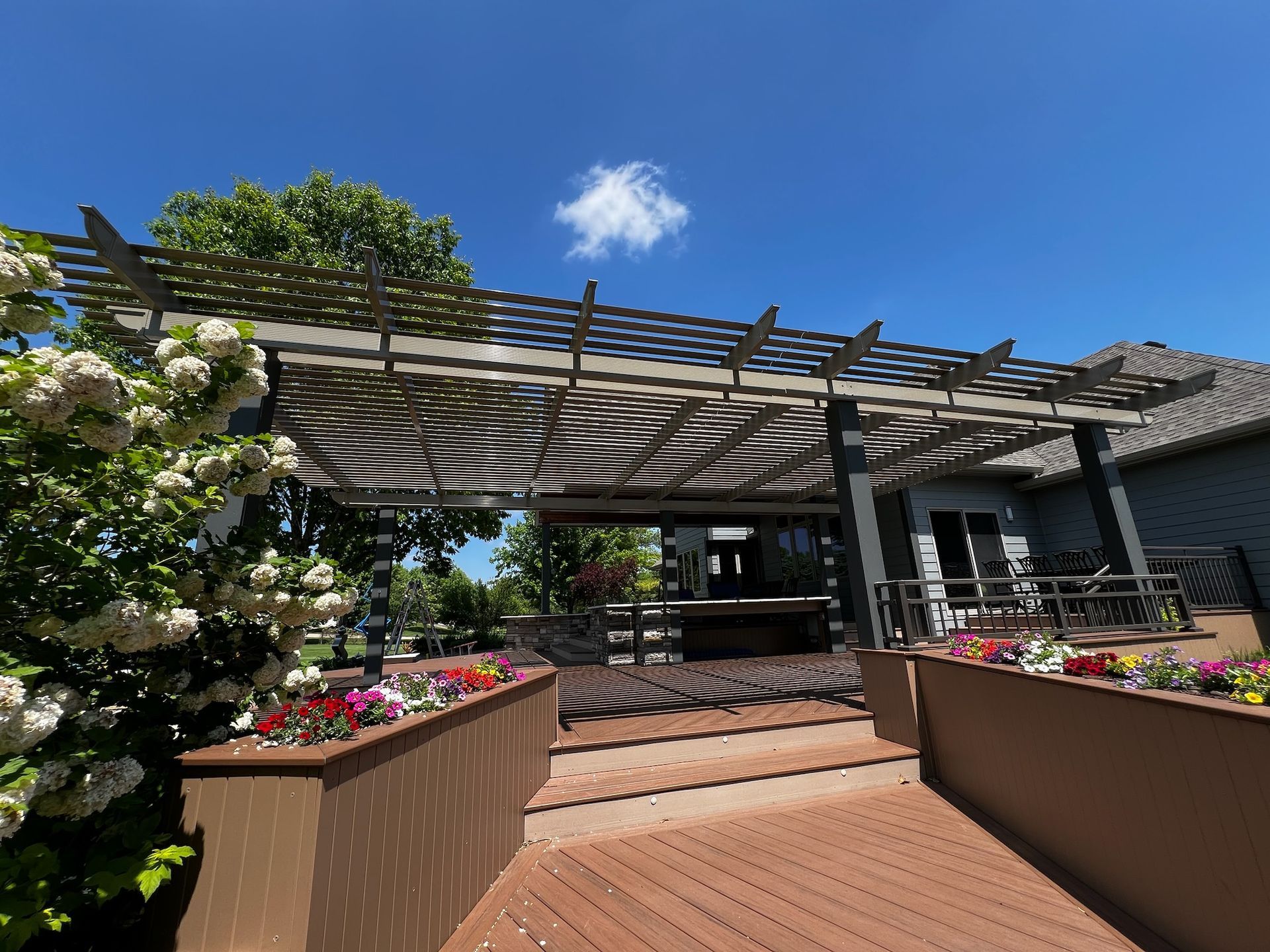 Patio garden entrance with flower planters, wooden pergola, and steps under a sunny blue sky