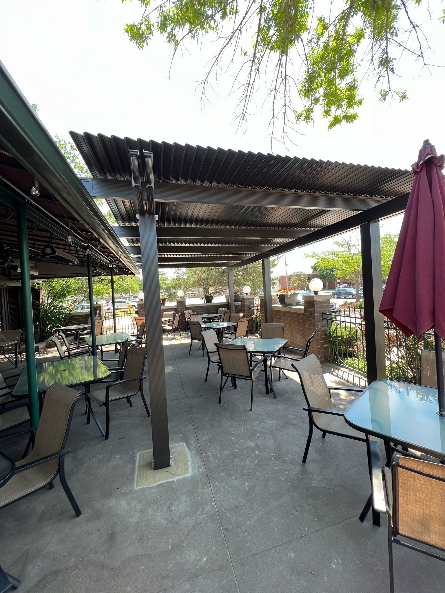 Outdoor café patio with metal tables and chairs under a corrugated roof, shaded by trees