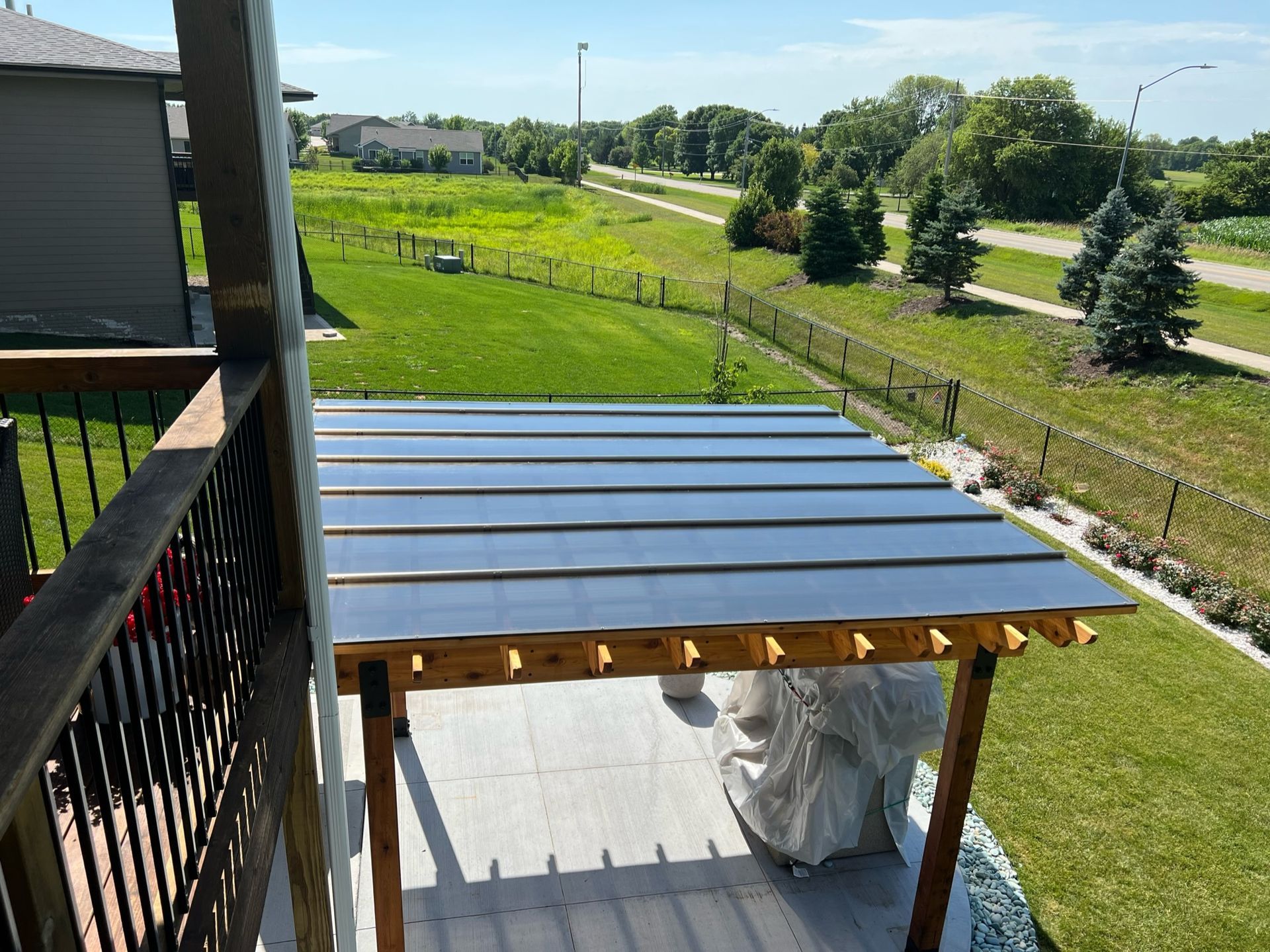 Covered patio with a metal roof beside a grassy yard and road on a sunny day