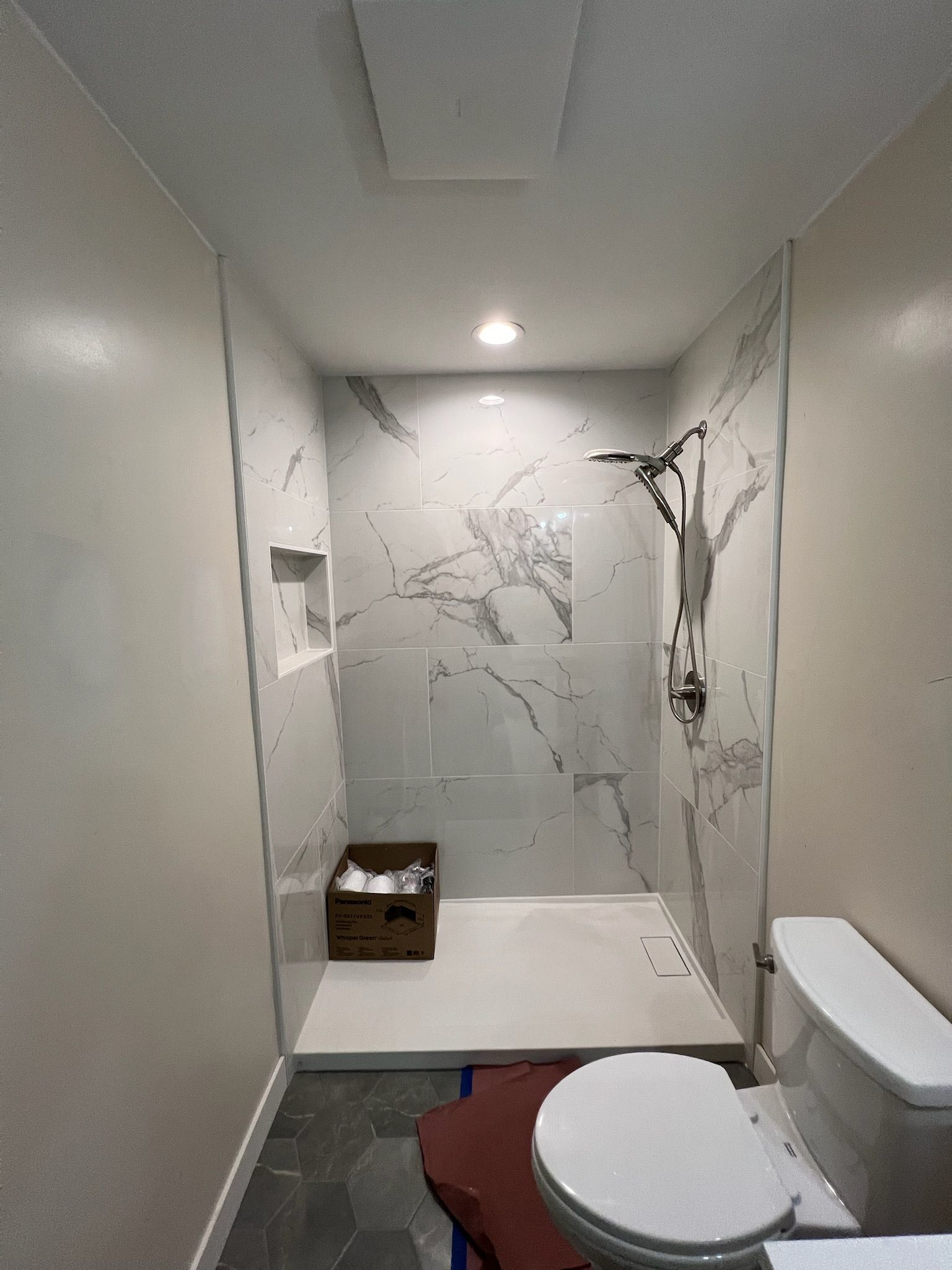 Small bathroom with white toilet, red bath mat, and damaged gray shower wall.