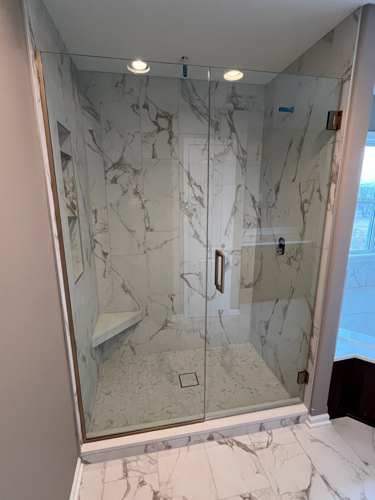 Marble-tiled glass shower stall with chrome fixtures and lighted ceiling in a bathroom
