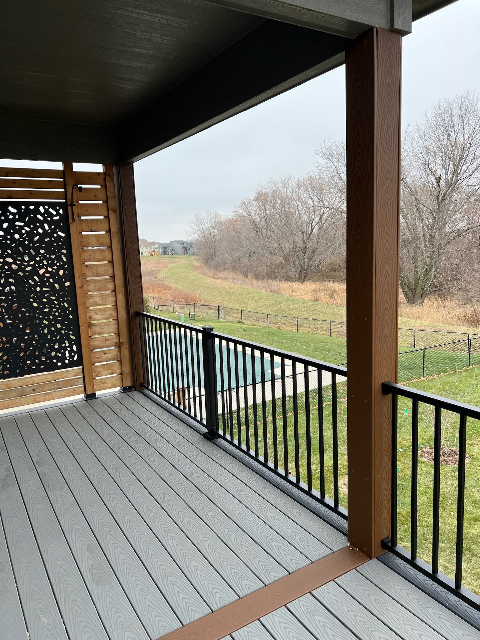 Covered porch with gray decking, black railing, and a view of grassy fields and bare trees beyond.