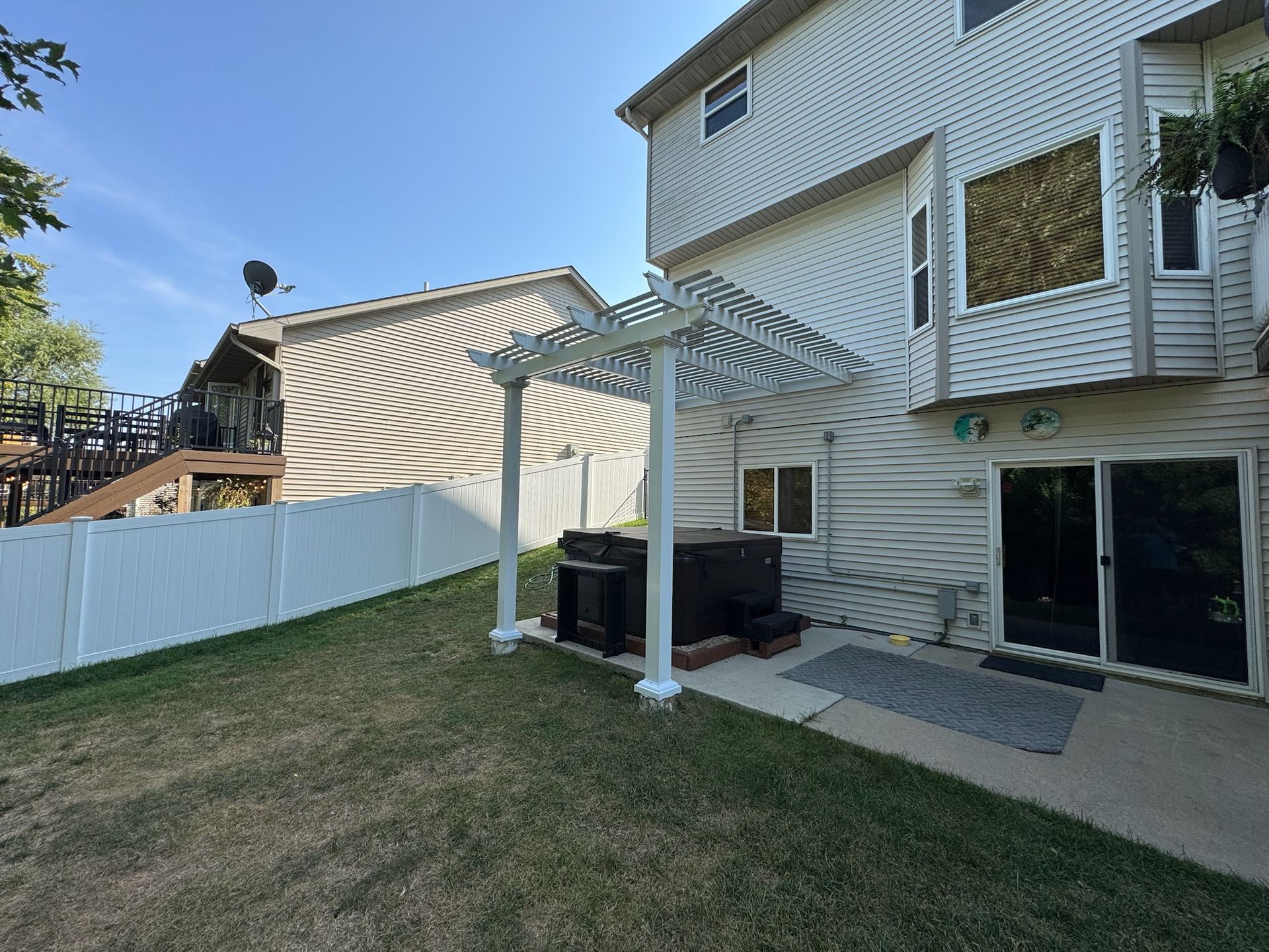 Backyard patio with white pergola, grill, fence, and a house exterior in sunlight