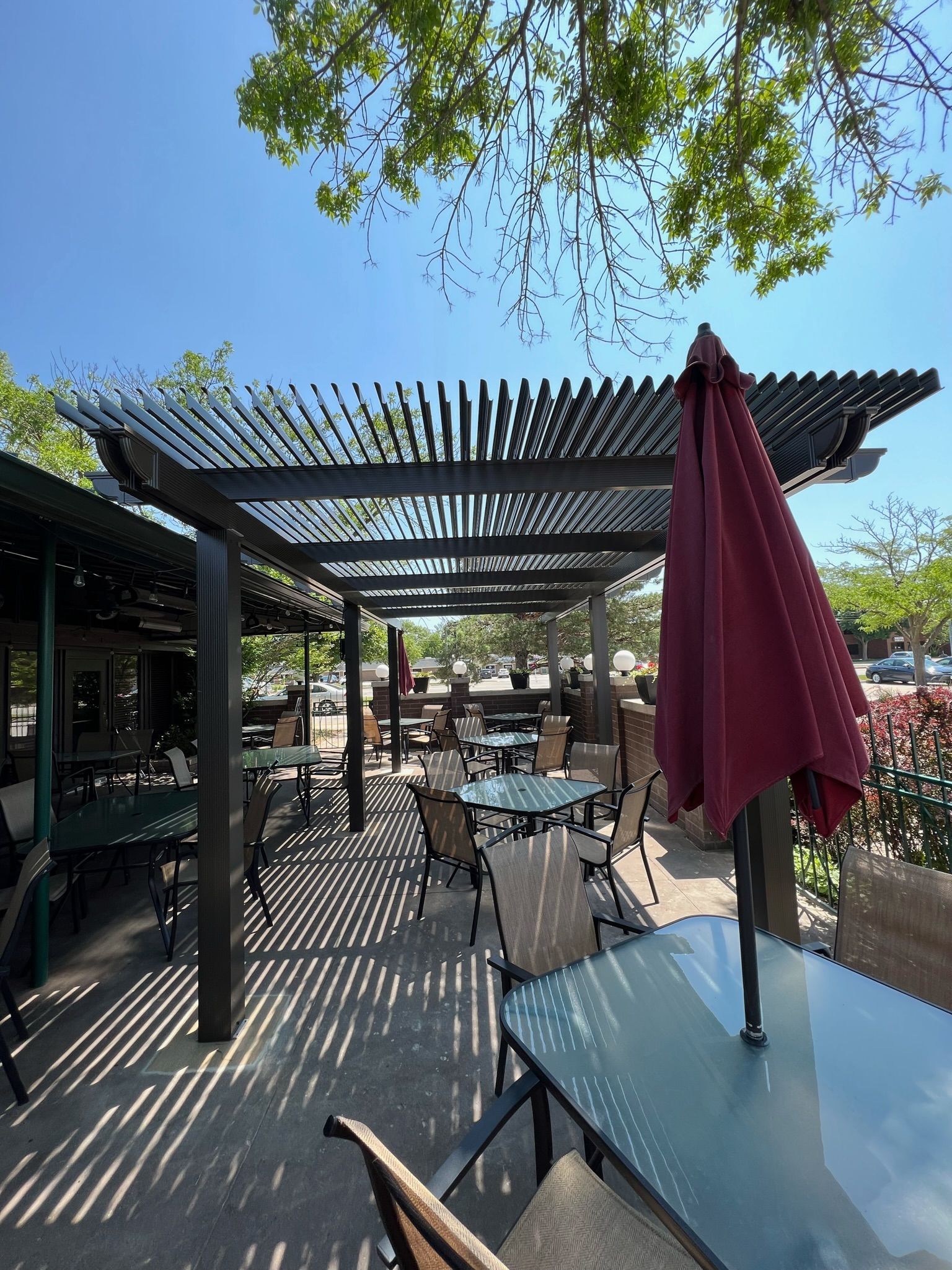 Outdoor café patio with black pergola, tables, and a closed red umbrella under leafy trees