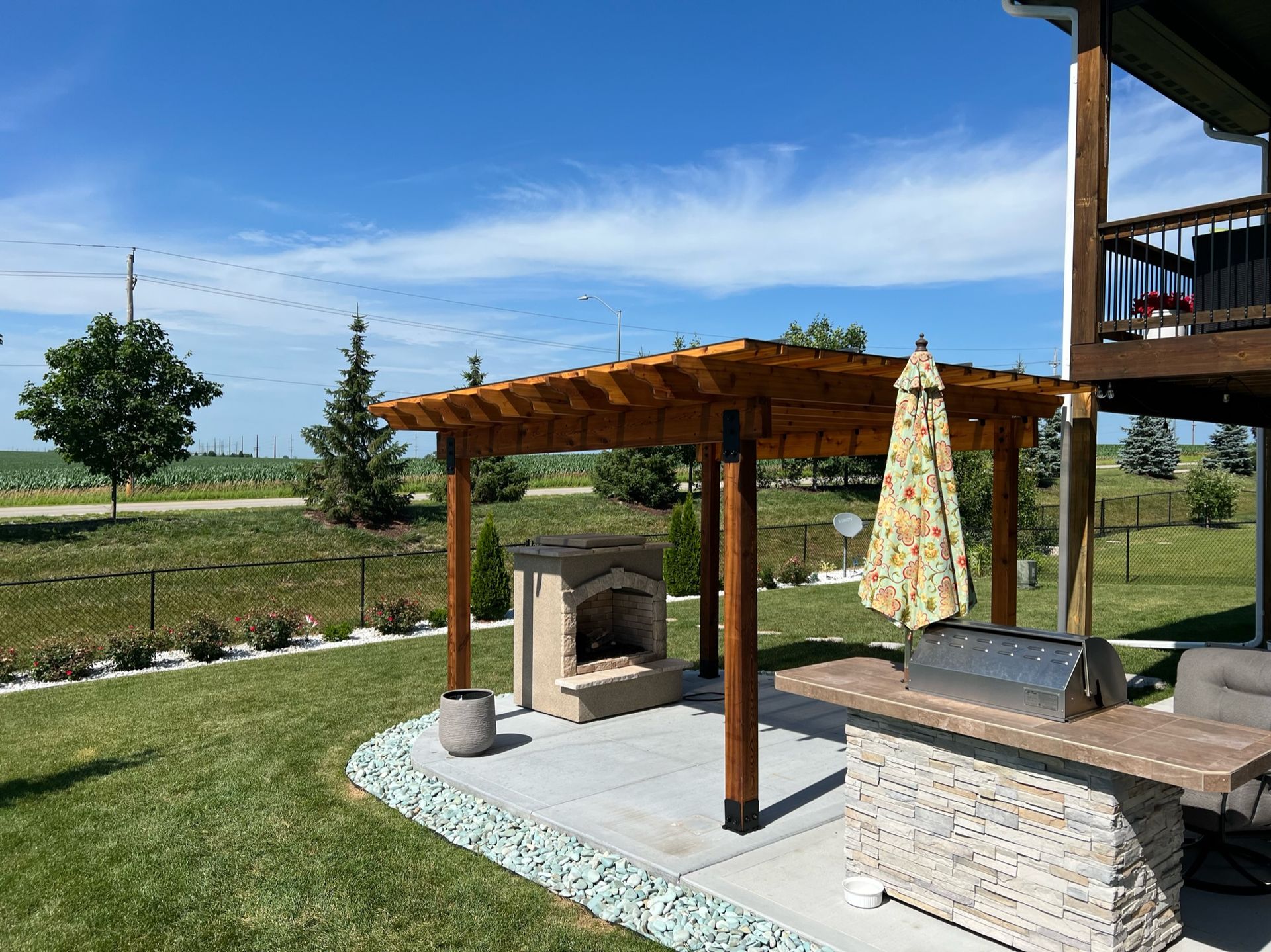Covered outdoor patio with wooden pergola, seating, umbrella, and grassy landscape under a blue sky