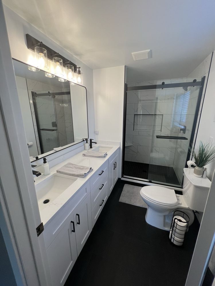 Modern, white bathroom with black accents, vanity, shower, toilet, and dark flooring.