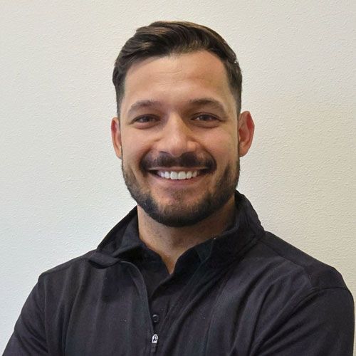Smiling man with dark hair and beard, wearing a black collared shirt, against a white background.