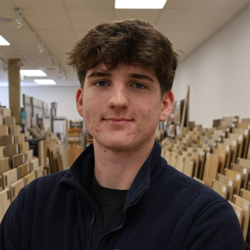 Young man in a navy jacket smiles in a flooring store, surrounded by wood samples.