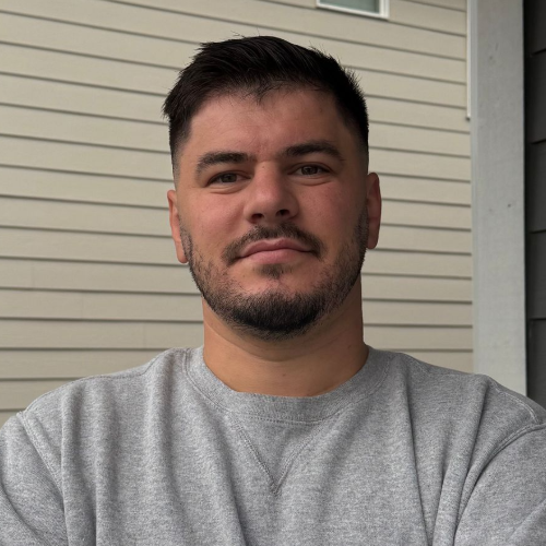 Man with crossed arms standing in front of a house, wearing a gray sweatshirt.