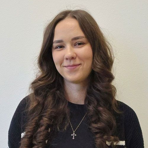Woman with long wavy brown hair smiles, wearing black sweater and cross necklace.