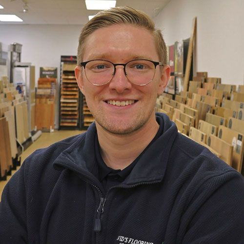 Man with glasses smiles in flooring store, surrounded by wood samples.