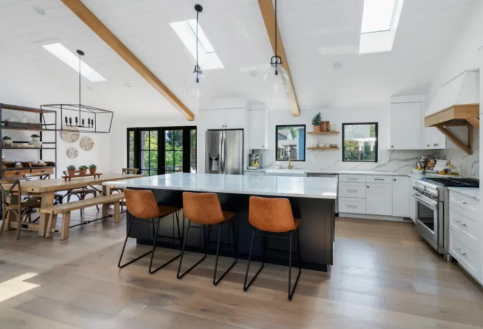 Modern kitchen with a large island, dining table, and skylights. Black island with brown stools and white cabinets.