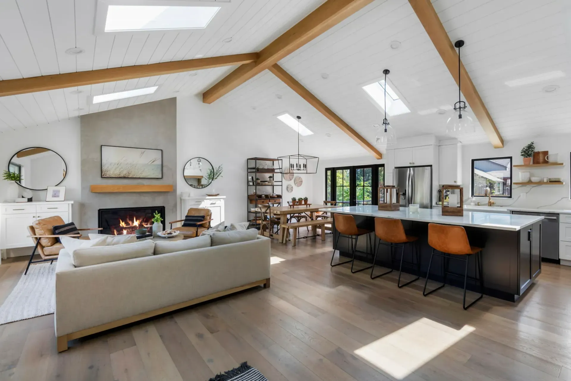 Spacious, open-plan living room with fireplace, kitchen island, and dining area; light wood floors, white walls, and exposed beams.