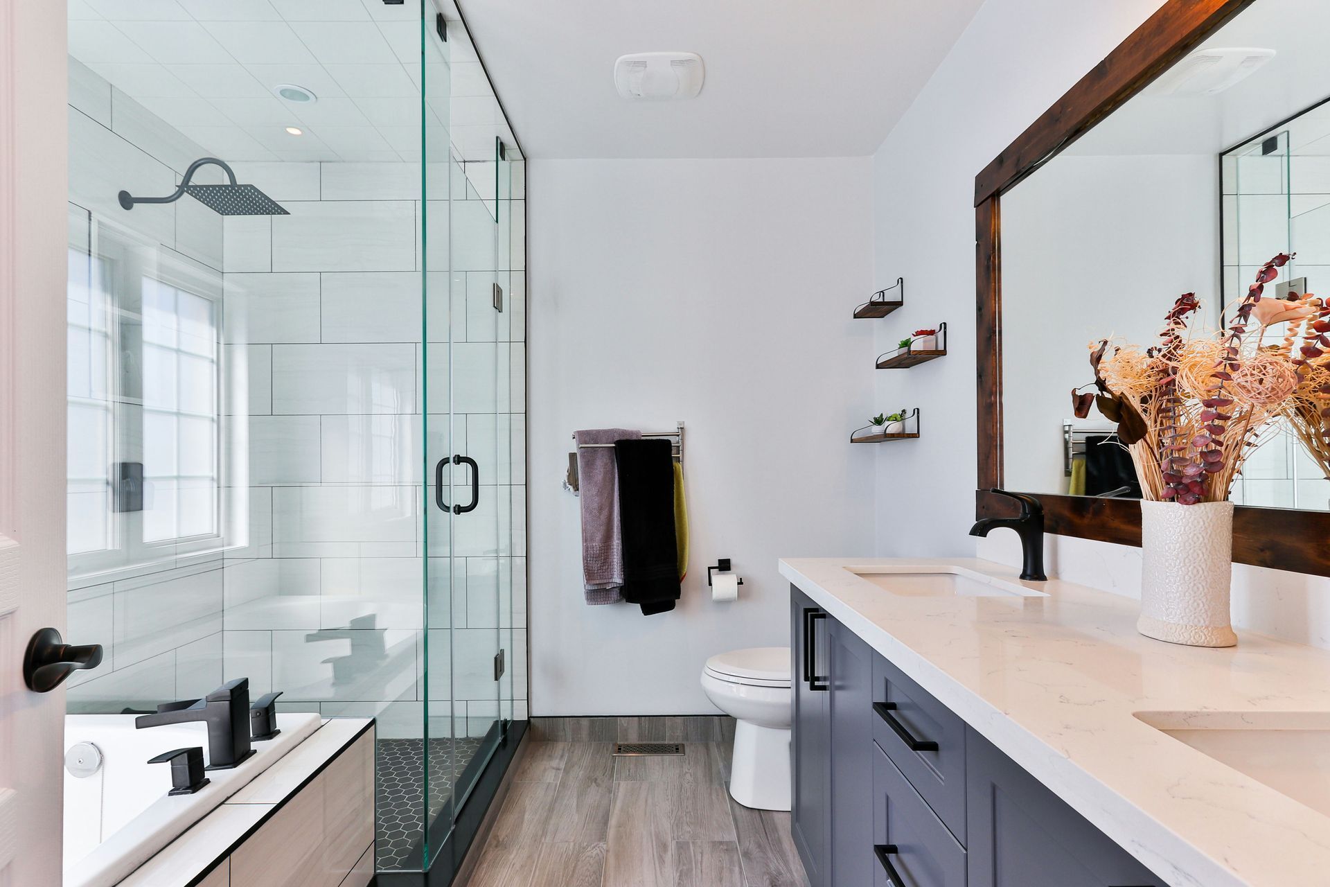 Modern bathroom with glass shower, double vanity, white walls, and wood-look flooring.