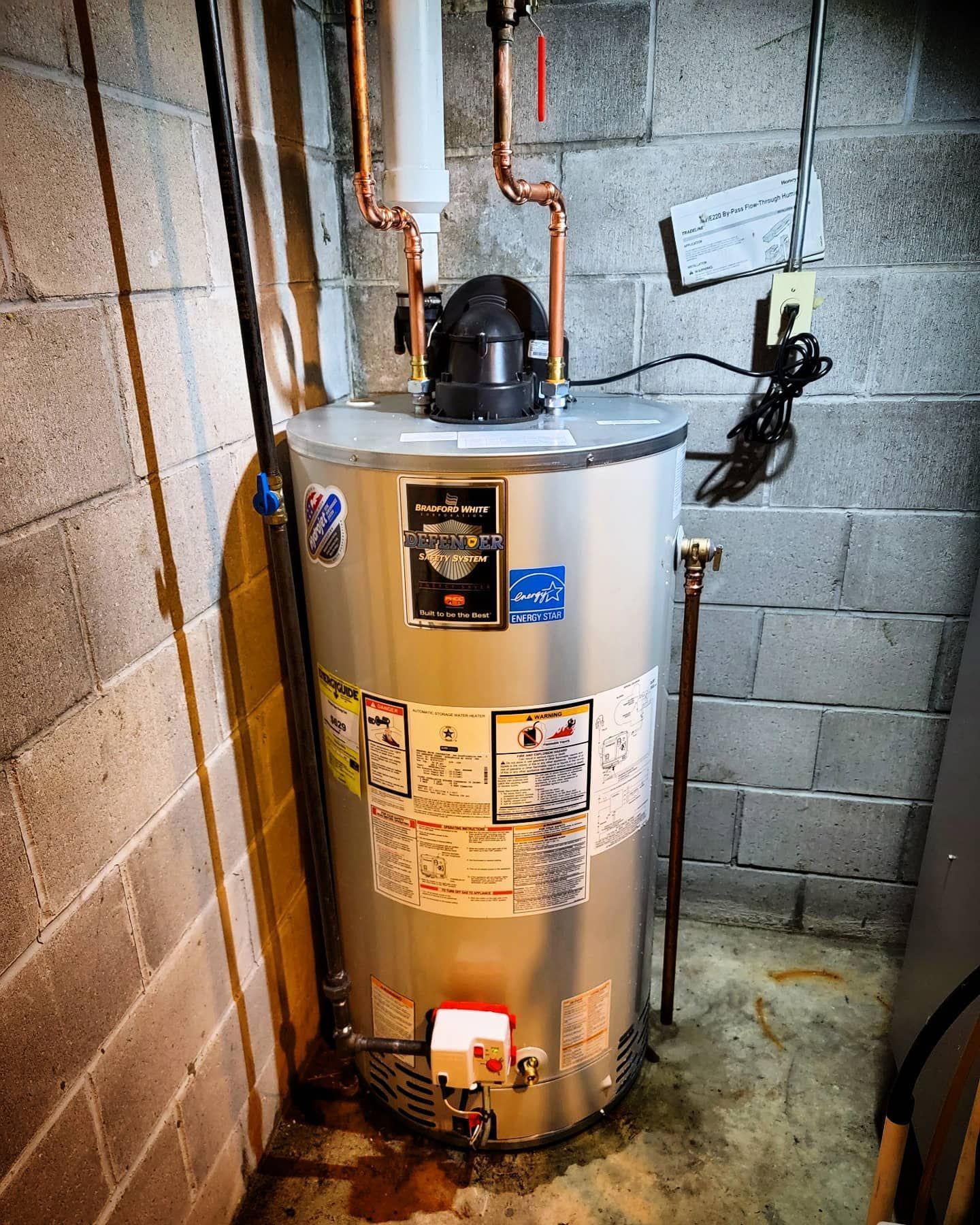 A water heater is sitting in a basement next to a brick wall.