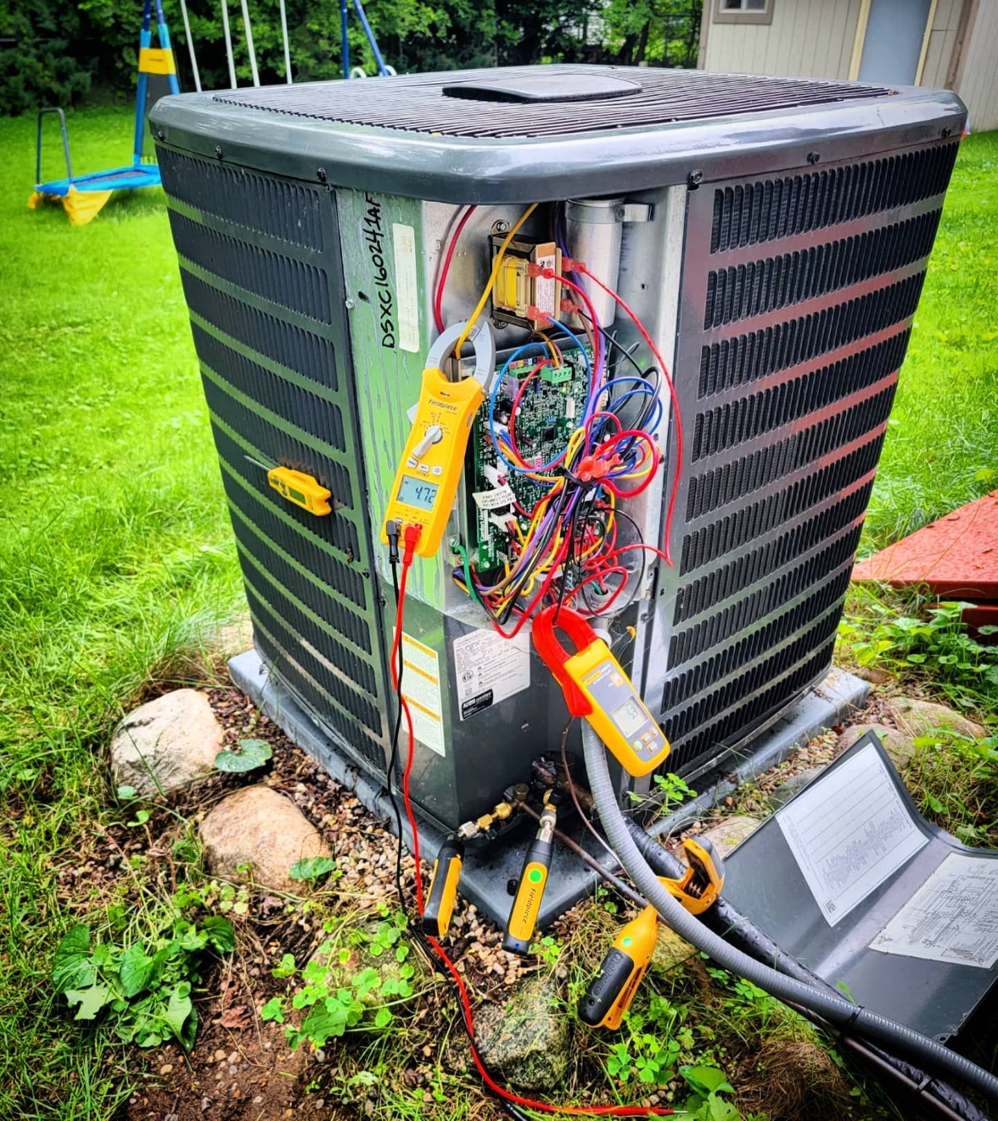 An air conditioner is being repaired in a yard with a bunch of wires coming out of it.