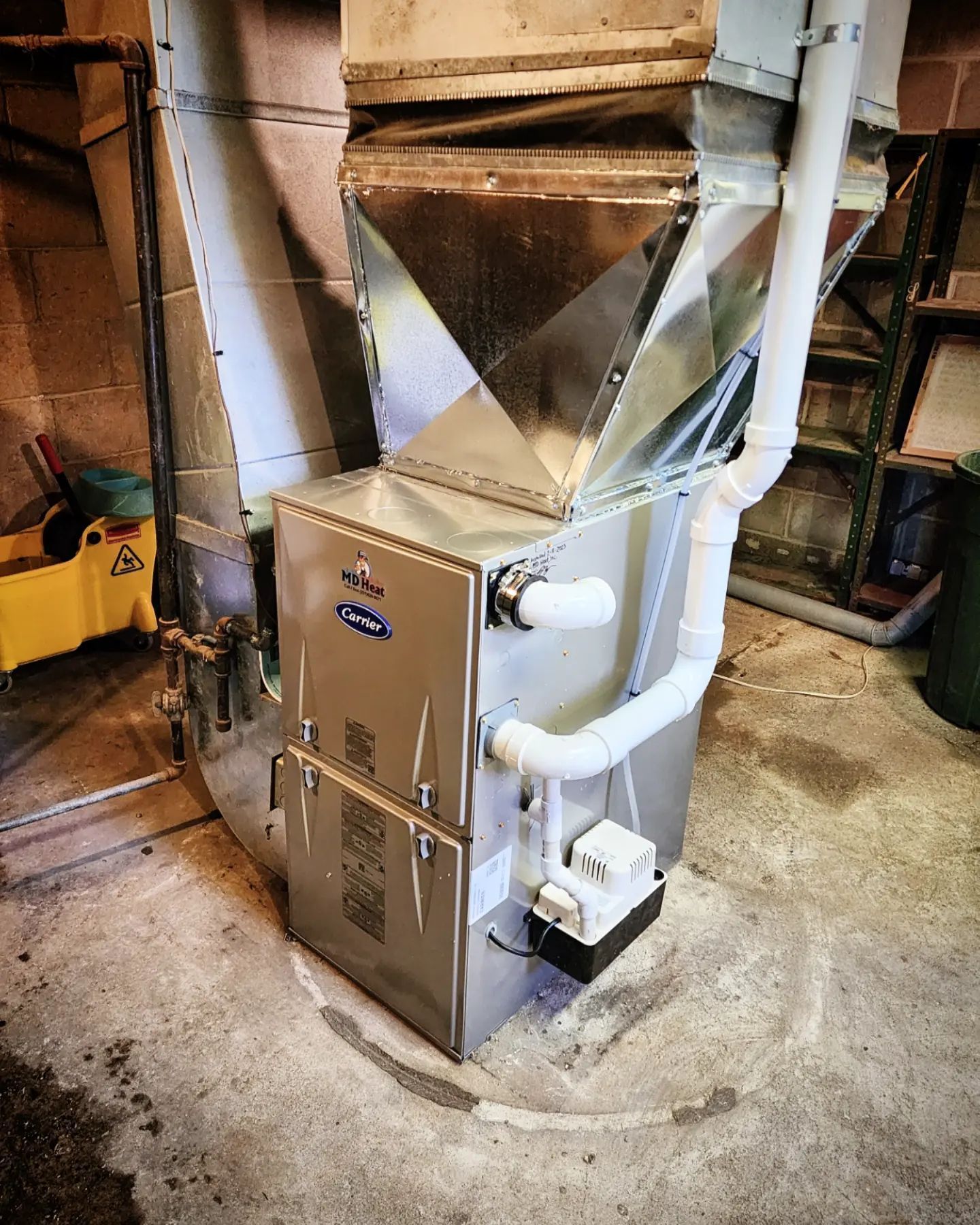 A stainless steel furnace is sitting in a basement next to a yellow mop.