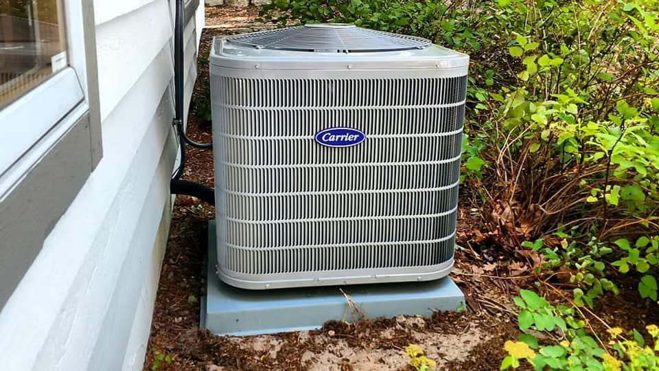A carrier air conditioner is sitting on the side of a house next to a window.