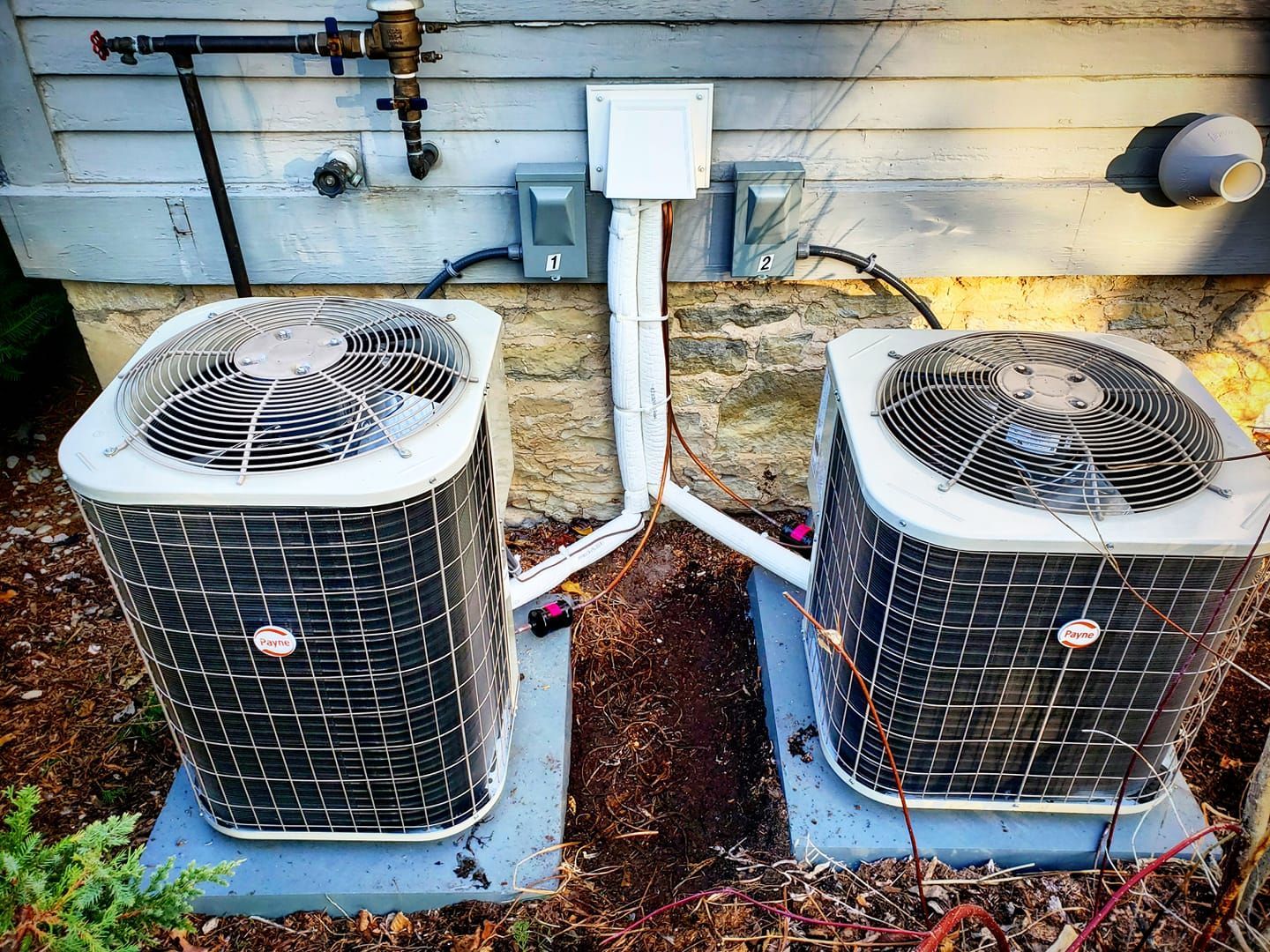 Two air conditioners are sitting next to each other on the side of a house.