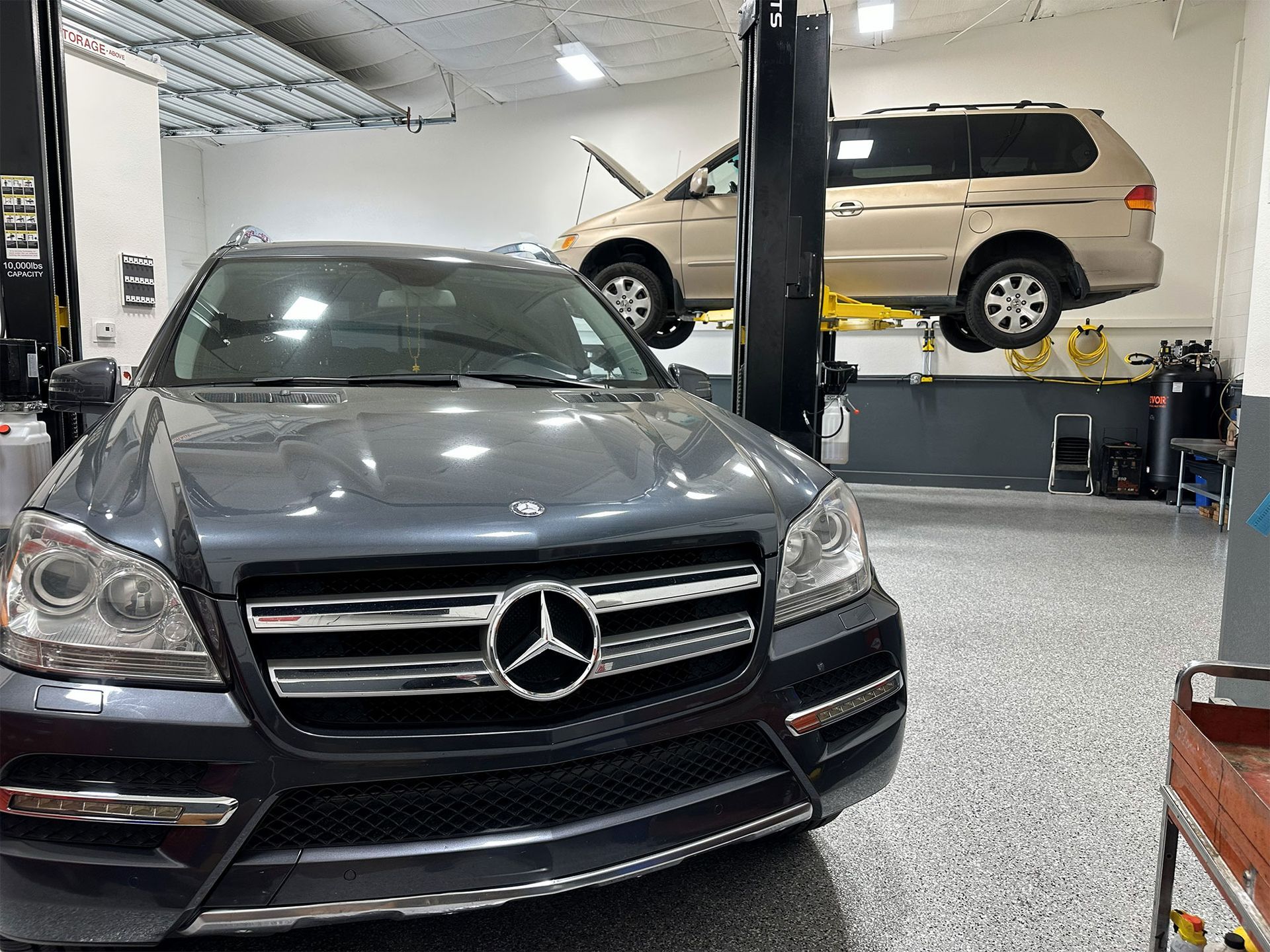 Gray Mercedes SUV in an auto repair shop with a van lifted on a hoist in the background.