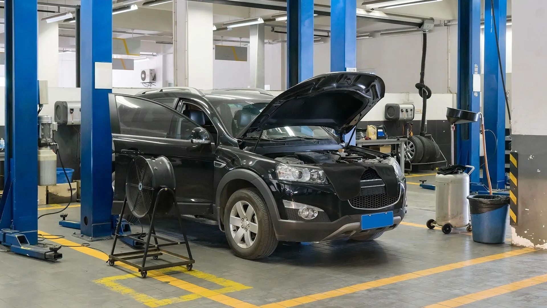 Black SUV in a mechanic shop with hood open, raised on a lift, blue support pillars, tools visible.