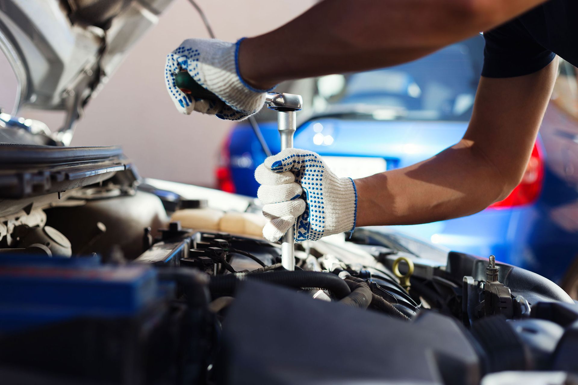 Mechanic's hands in gloves using a wrench on a car engine. Bright outdoor setting, open hood, blue car in background.