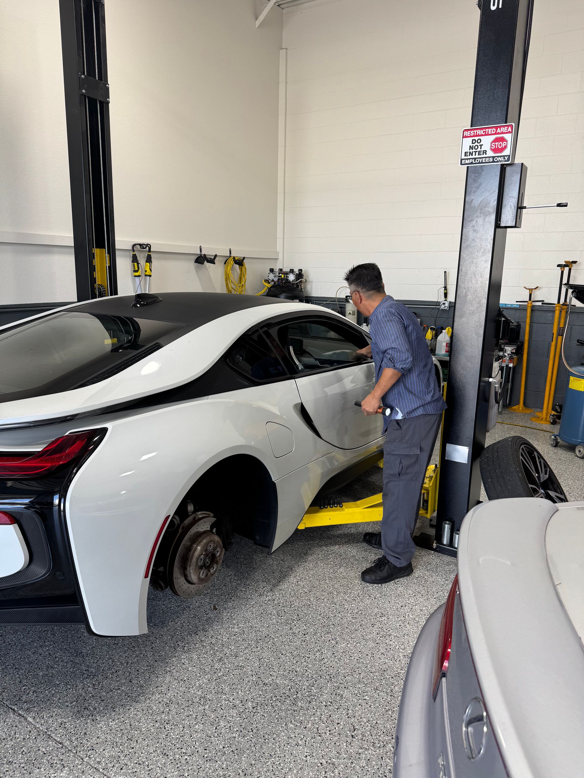 Mechanic working on a white BMW i8 in a garage; car on a lift with one tire removed.