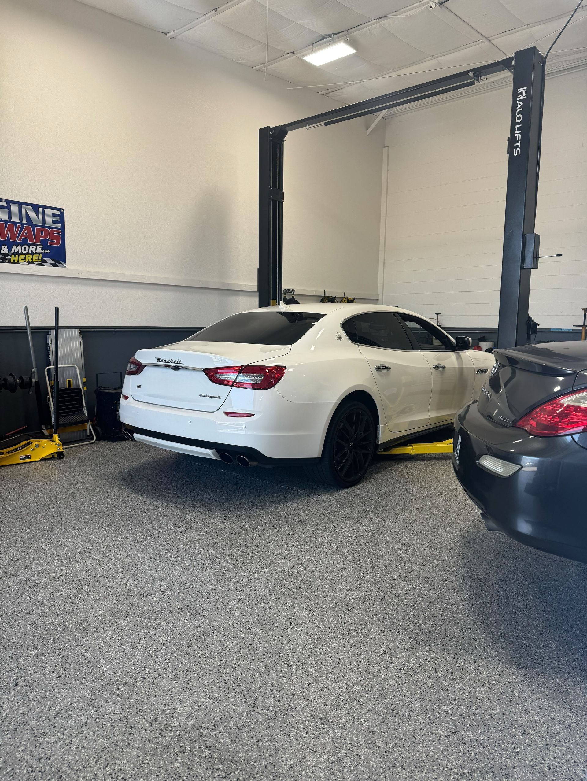 White car on a lift in a garage, next to a dark car.