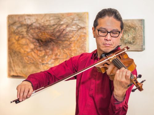 Violinist in a red shirt playing in front of abstract artwork, focused expression