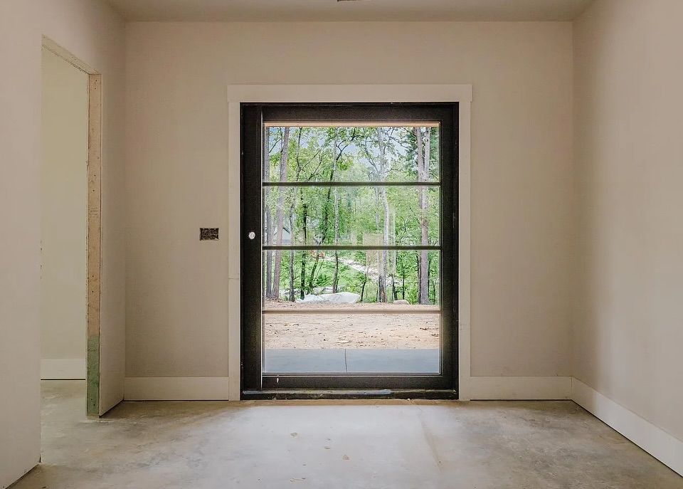 Interior view of a room with a black-framed glass door overlooking trees. Light-colored walls, concrete floor.