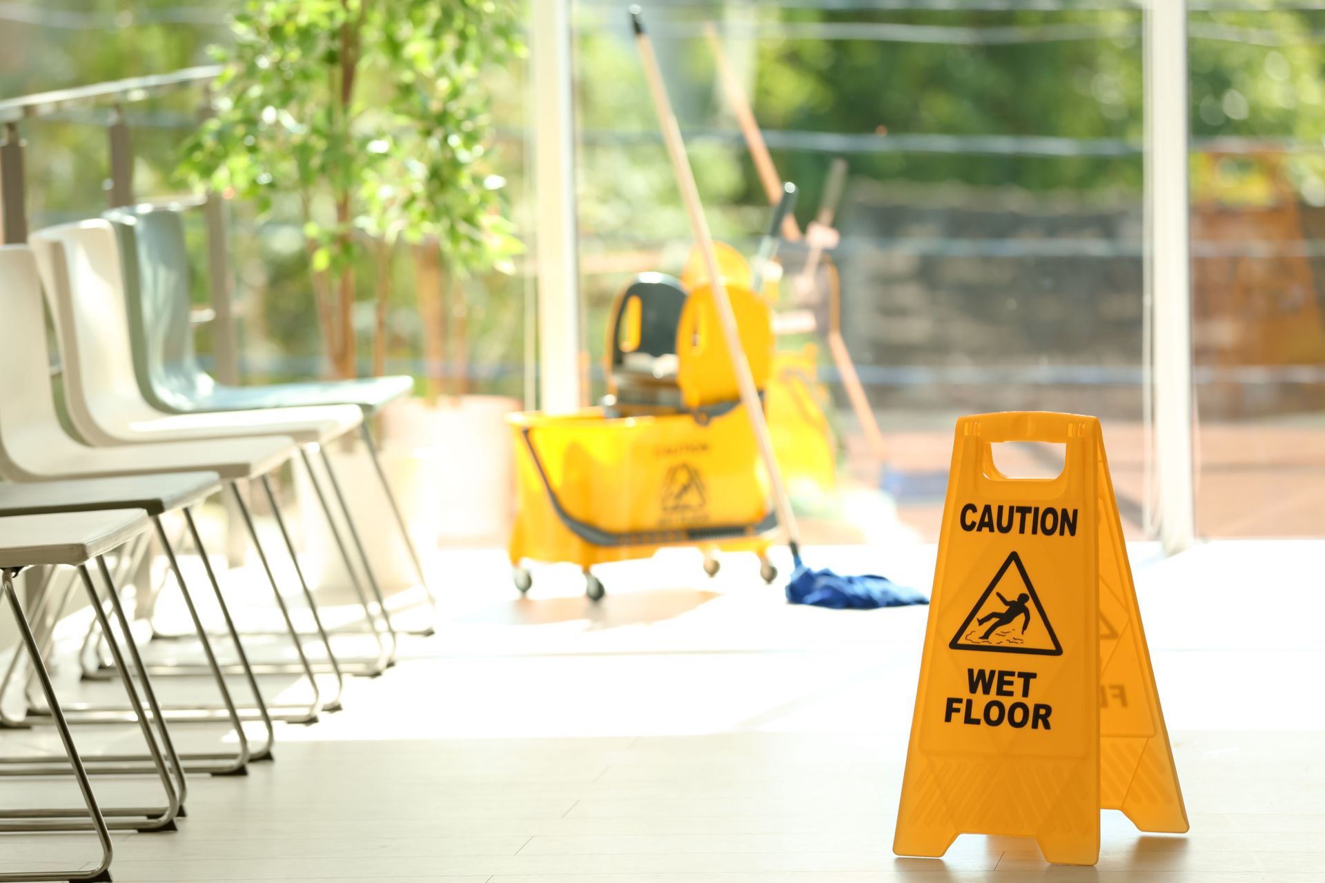 Caution wet floor sign beside a mop and bucket in a bright hallway with chairs and sunlight