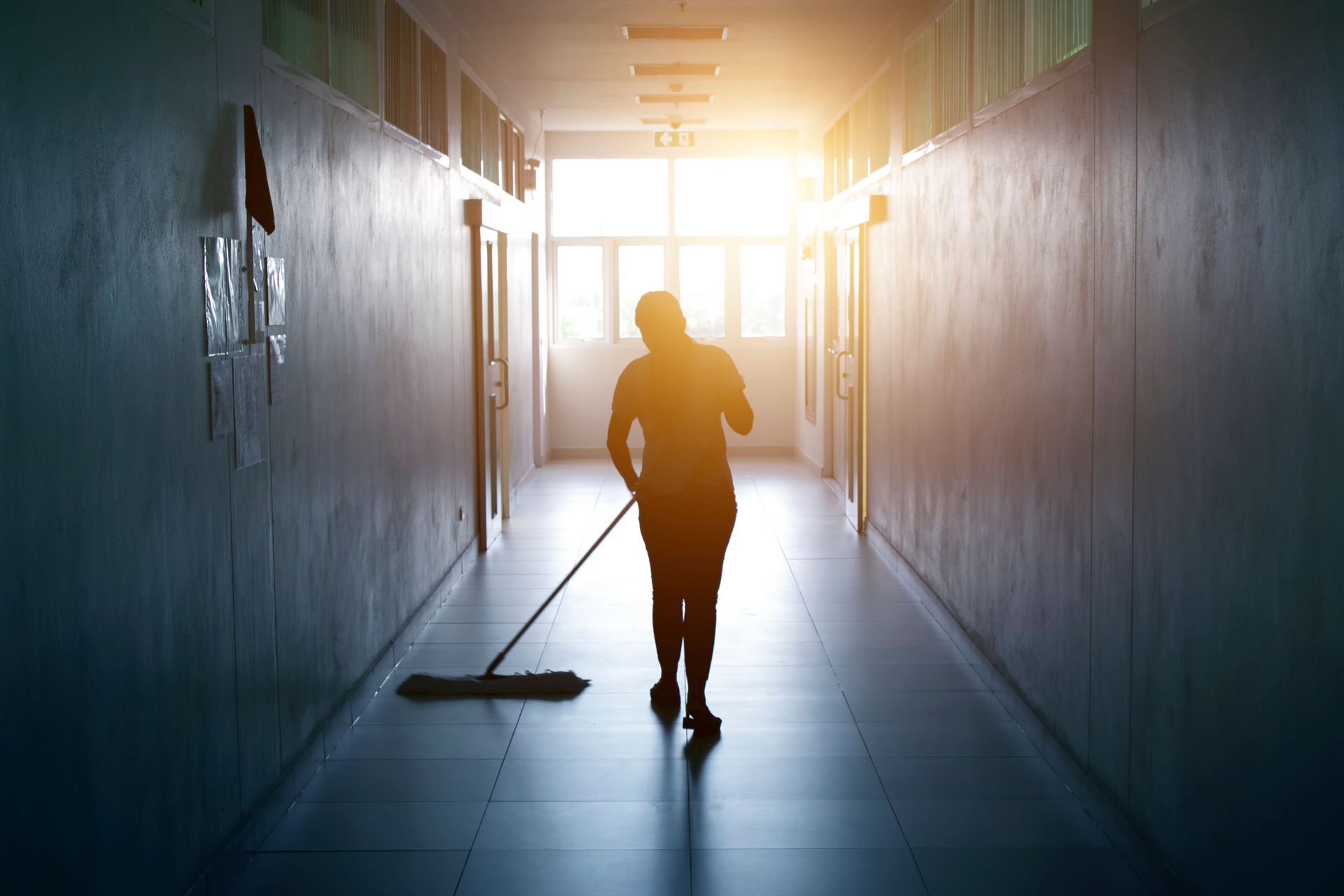 Silhouette of a person running down a bright hallway, mopping the floor toward the light.