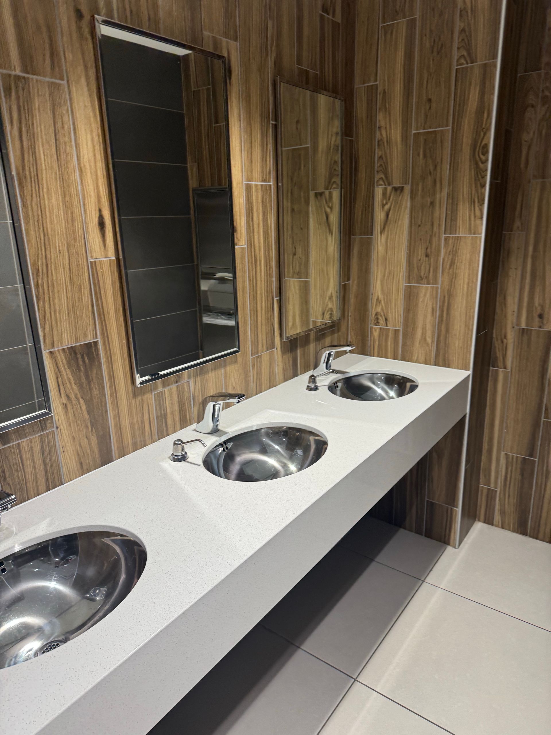 Triple stainless-steel sinks on a white counter in a wood-paneled restroom with mirrors.