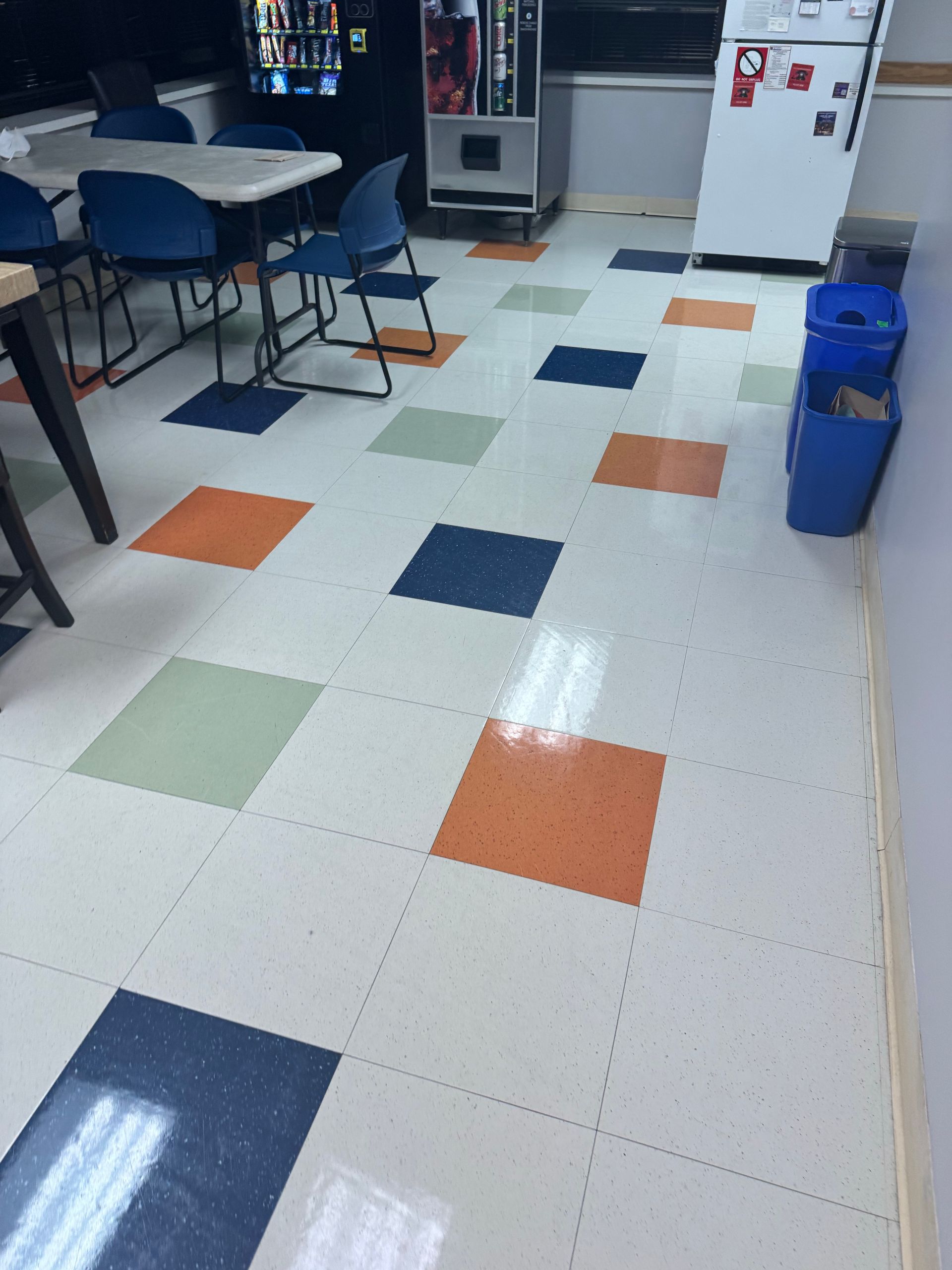 Indoor room with white tile floor and colorful square accents, tables and chairs on the left, bins on the right.