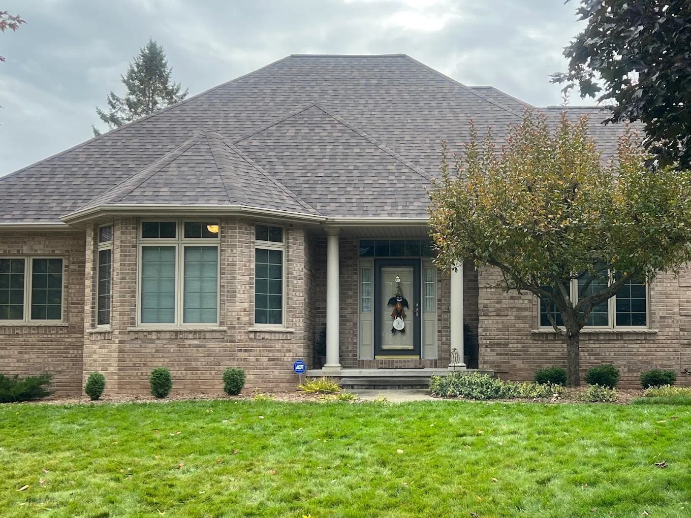 Brick house with brown roof, front porch with columns, and small tree on a green lawn.