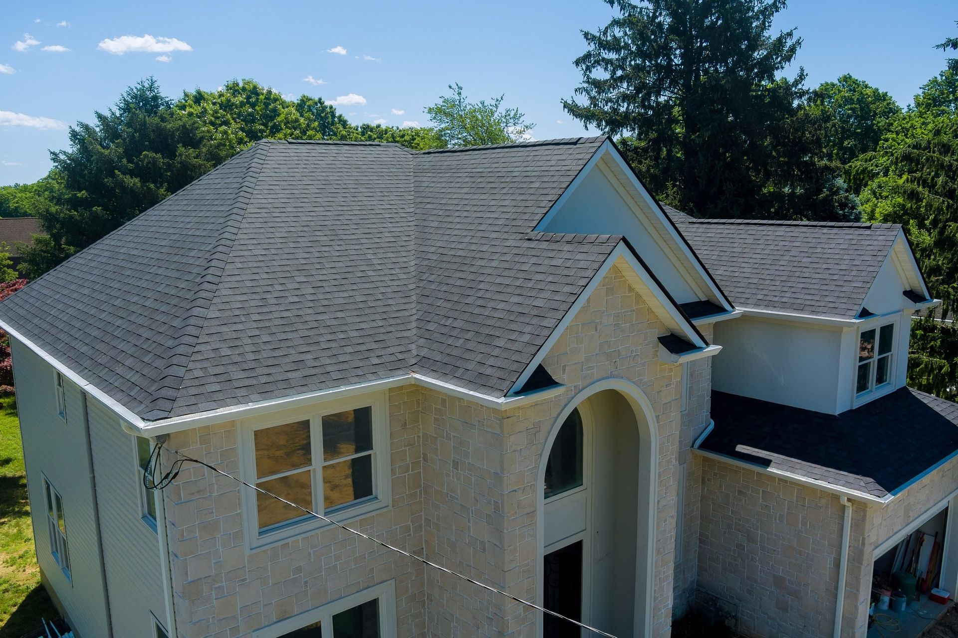 House with dark gray roof, beige exterior, and surrounding green trees under a blue sky.