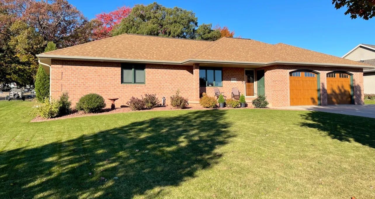 Brick ranch-style house with brown roof and garage doors, green lawn, and shrubs.