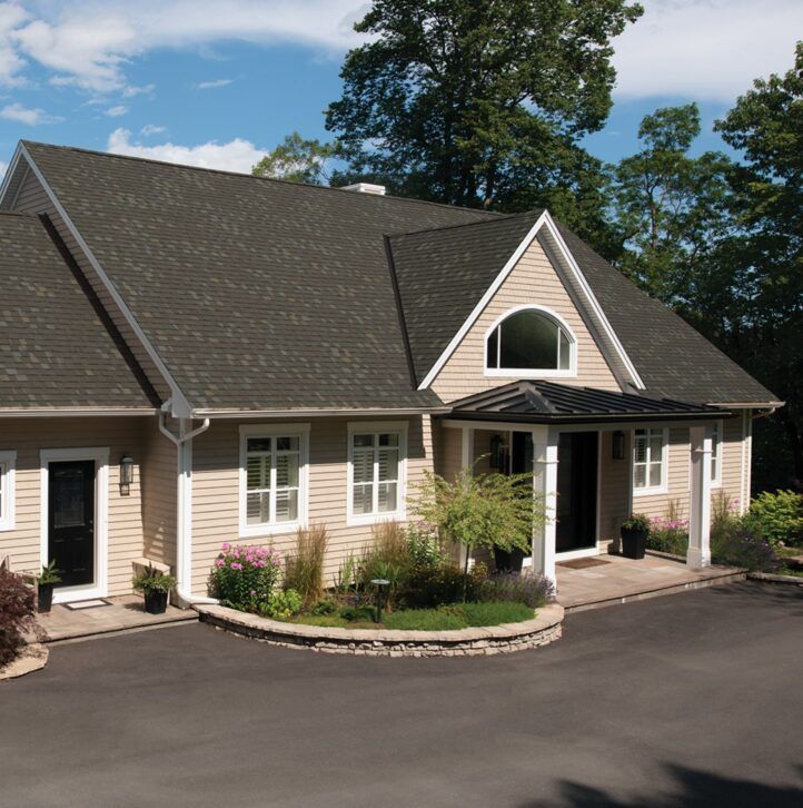 Tan house with dark roof and black front door. Black asphalt driveway. Green trees in background.