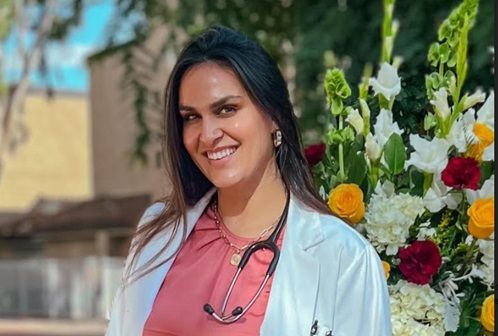 Katie Hammond, FNP-BC  in a lab coat with a stethoscope around her neck is smiling in front of flowers .