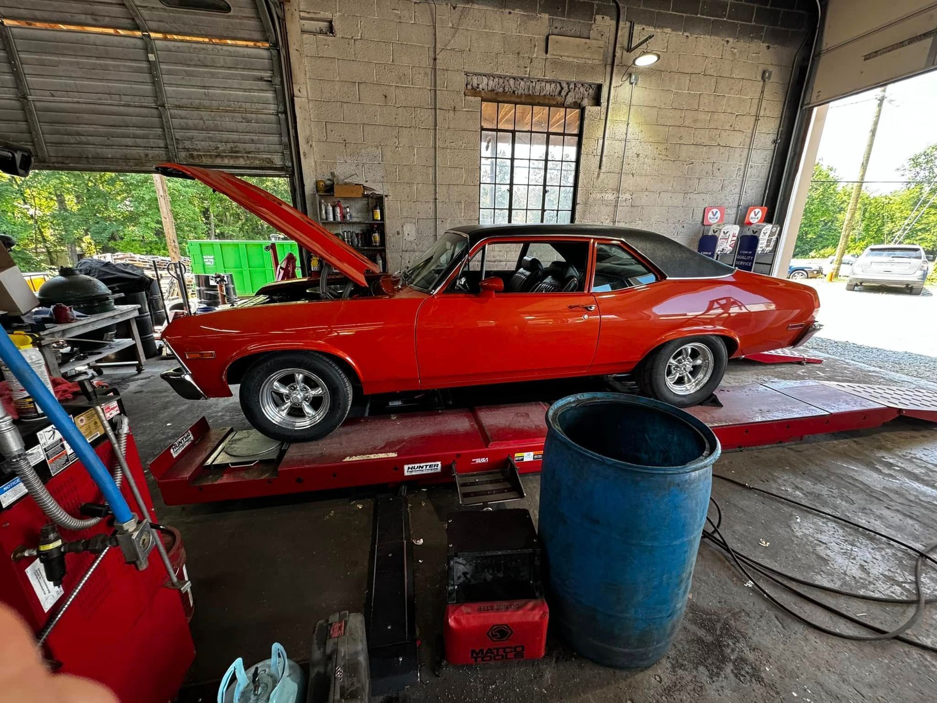 Red classic car on a lift with hood open in a garage, next to a blue barrel.